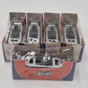 Puede incluir: Un conjunto de seis interruptores eléctricos en su embalaje original. Los interruptores son plateados y negros, y el embalaje es una mezcla de azul, rojo y blanco con el logotipo "EAGLE".