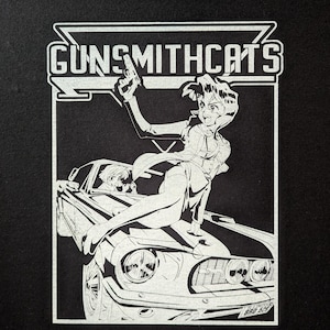 May include: Black and white graphic featuring the text "GUNSMITHCATS" above an illustration of a woman holding a gun, posed on the hood of a classic car. Another person is visible in the driver's seat. The image has a retro anime style.