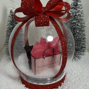 May include: A clear glass snow globe with a red glitter rim and a red glitter bow on top. Inside the globe is a pink gift bag with the text "VICTORIA'S SECRET" and white faux snow.