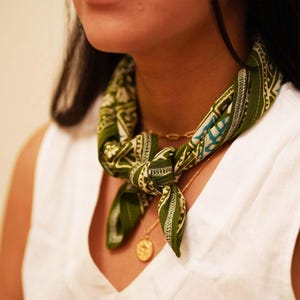 May include: A square bandana featuring an olive green base with a white and teal patterned print, tied in a knot around the neck. The fabric displays a repeating geometric and floral design with visible white stitching along the finished edges.