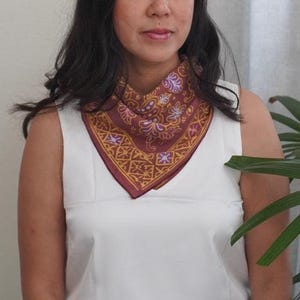 May include: A burgundy scarf with a detailed floral and geometric pattern in gold and purple. The scarf is worn around the neck, creating a triangular shape. The background is a white top and a green plant.