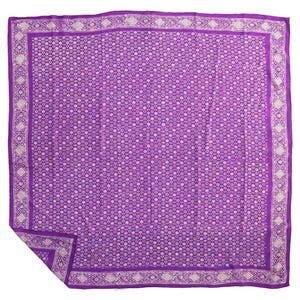 May include: A square purple scarf featuring a repeating white geometric pattern across the center and a decorative border with diamond shapes. The fabric has a smooth texture and is folded at one corner to reveal the underside of the material.