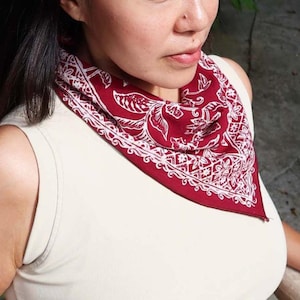 May include: A burgundy bandana with a white floral and paisley design is worn around the neck. The bandana is folded into a triangle shape. The person is wearing a cream-colored tank top.