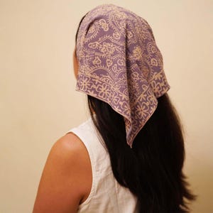 May include: A lavender bandana with a beige floral pattern is worn on the head. The square scarf is folded and tied at the back of the head, with the corners pointing down. The bandana is made of a soft fabric and is a stylish accessory.