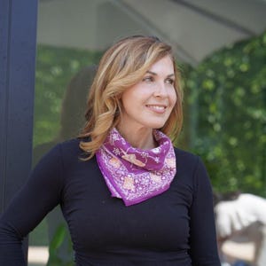 May include: A purple bandana-style scarf with a floral and geometric pattern. The scarf is worn around the neck. The scarf has a square shape and is made of a soft fabric. The scarf is a vibrant accessory.