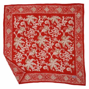 May include: A red square scarf with a floral pattern in white and tan. The scarf has a decorative border with geometric shapes. The floral design features large flowers and vines. The scarf is made of a soft fabric.