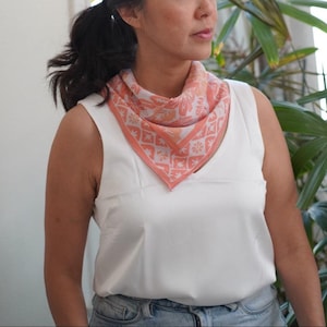 May include: A square scarf featuring a coral orange and white floral and geometric pattern, folded into a triangle and worn around the neck. The scarf has a solid coral orange border and a repeating design of stylized flowers and star-like shapes.