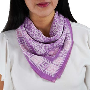 May include: A square scarf in shades of purple and white, featuring a repeating floral and geometric pattern. The scarf is draped around the neck, showcasing its intricate design and color palette. The scarf's border has a Greek key design.