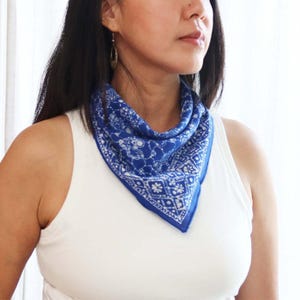 May include: A triangular bandana featuring a vibrant blue background with a white intricate floral and geometric pattern. The fabric is folded and tied around the neck, displaying a repeating motif of small blossoms and diamond shapes across the surface.