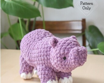 May include: A handmade, lavender-colored crochet hippo with white accents on its feet. The plush toy is displayed on a light-colored surface, with a leafy green plant in the background. The image includes the text "Pattern Only".