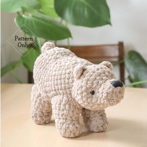 May include: A light brown, crocheted bear with a black nose and small ears. The bear is standing on a light-colored surface. The text "Pattern Only" is visible in the background.