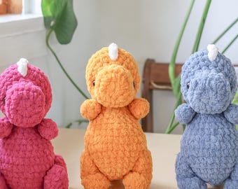 May include: Three handmade crochet dinosaur plushies in a row. The dinosaurs are in shades of pink, orange, and blue. Each has a white horn and small arms and legs. The plushies are sitting on a light brown surface.