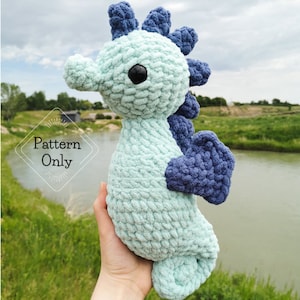 May include: A handmade, plush seahorse in shades of blue and teal. The seahorse has a textured, soft appearance, with a dark eye and a curled tail. The text "Pattern Only" is visible in the image. This item is a crochet pattern.