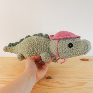 Op de afbeelding: Een groene gehaakte alligator die een roze gehaakte hoed draagt. De alligator wordt vastgehouden door een hand.