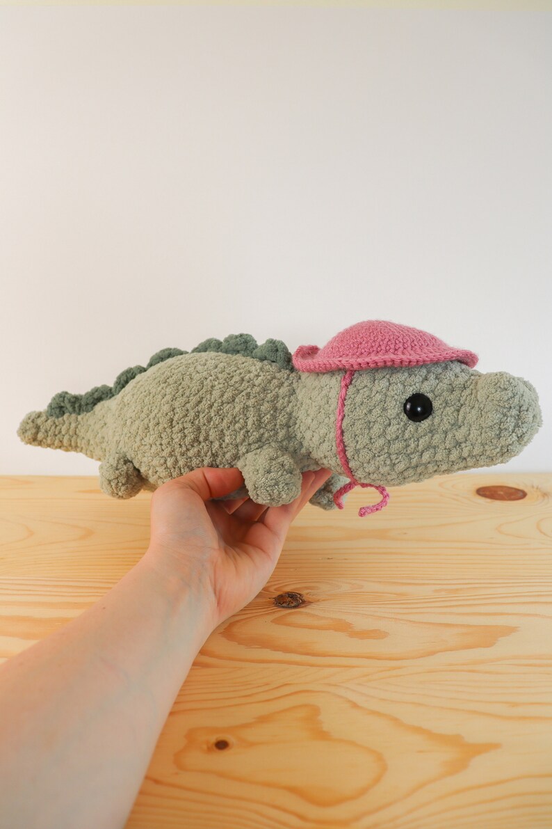 Op de afbeelding: Een groene gehaakte alligator die een roze gehaakte hoed draagt. De alligator wordt vastgehouden door een hand.