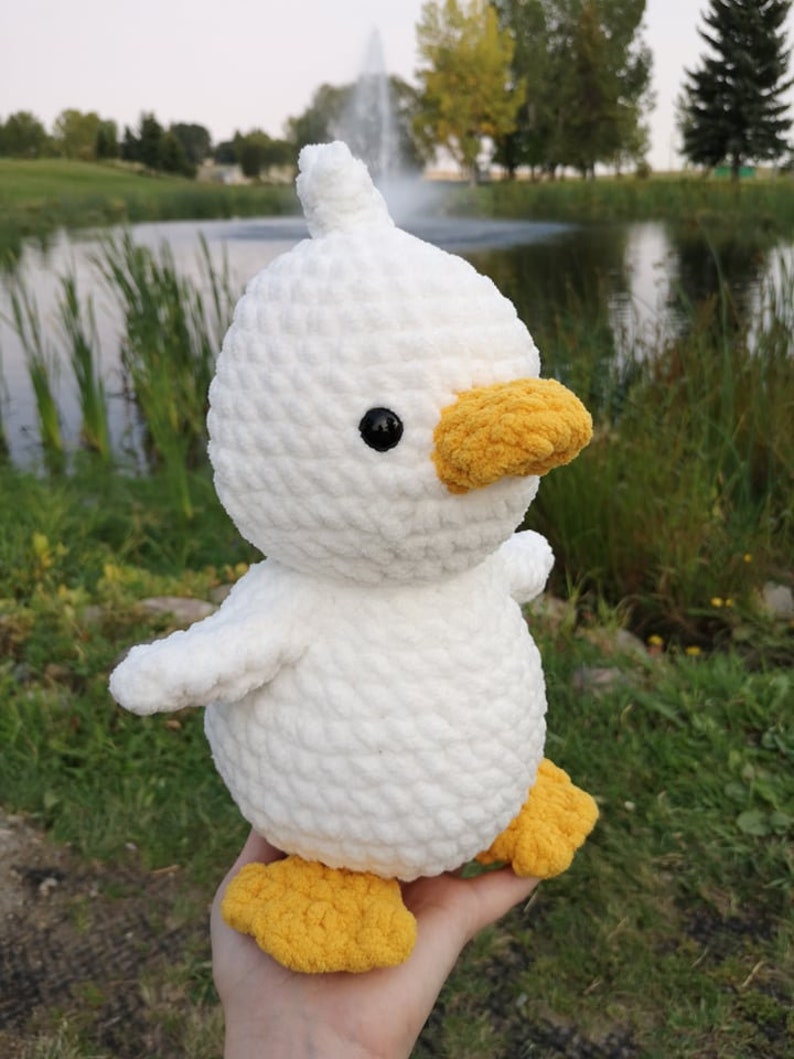 May include: White crocheted duck plush toy with yellow beak and feet. The duck has black eyes and a white fluffy body.