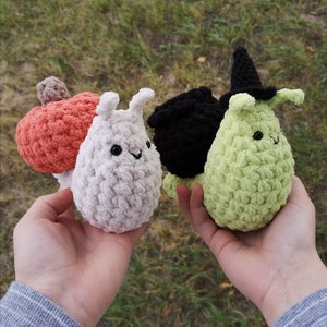 May include: Three crocheted snail plushies. The first snail is white with a pumpkin on its back. The second snail is black with a witch's cauldron on its back. The third snail is green with a witch's hat on its head.