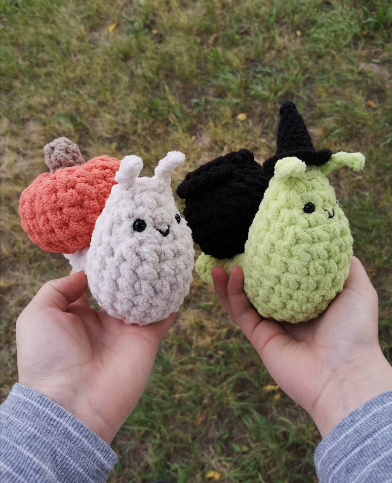 May include: Three crocheted snail plushies. The first snail is white with a pumpkin on its back. The second snail is black with a witch's cauldron on its back. The third snail is green with a witch's hat on its head.