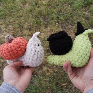 May include: Two crocheted snail plushies. One is white with a peach-colored pumpkin on its back. The other is green with a black witch's hat and cauldron.