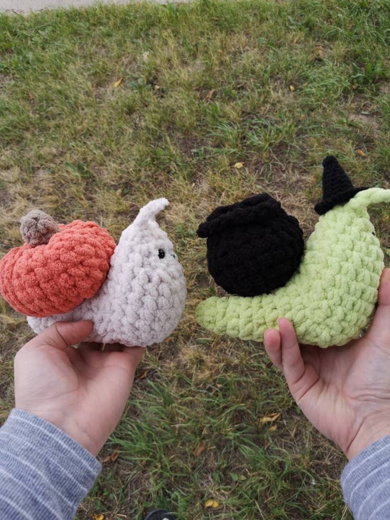 May include: Two crocheted snail plushies. One is white with a peach-colored pumpkin on its back. The other is green with a black witch's hat and cauldron.