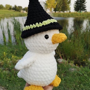 May include: A white crocheted duck wearing a black witch hat with a green trim. The duck has yellow feet and a yellow beak.