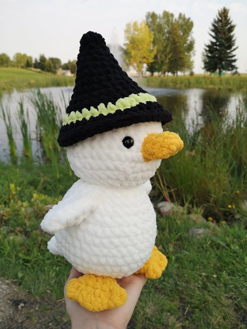 May include: A white crocheted duck wearing a black witch hat with a green trim. The duck has yellow feet and a yellow beak.