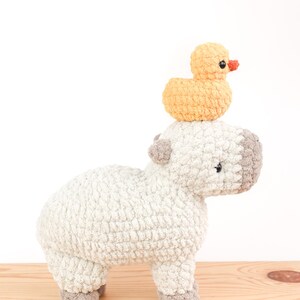 May include: A handmade crochet stuffed animal featuring a light gray capybara with gray accents and a small yellow duck perched on its head. The duck has an orange beak and black eyes. The toys are on a wooden surface.