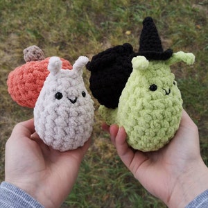 May include: Three crocheted plush toys in the shape of snails. The snails are white, green, and black. The green snail is wearing a black witch's hat.