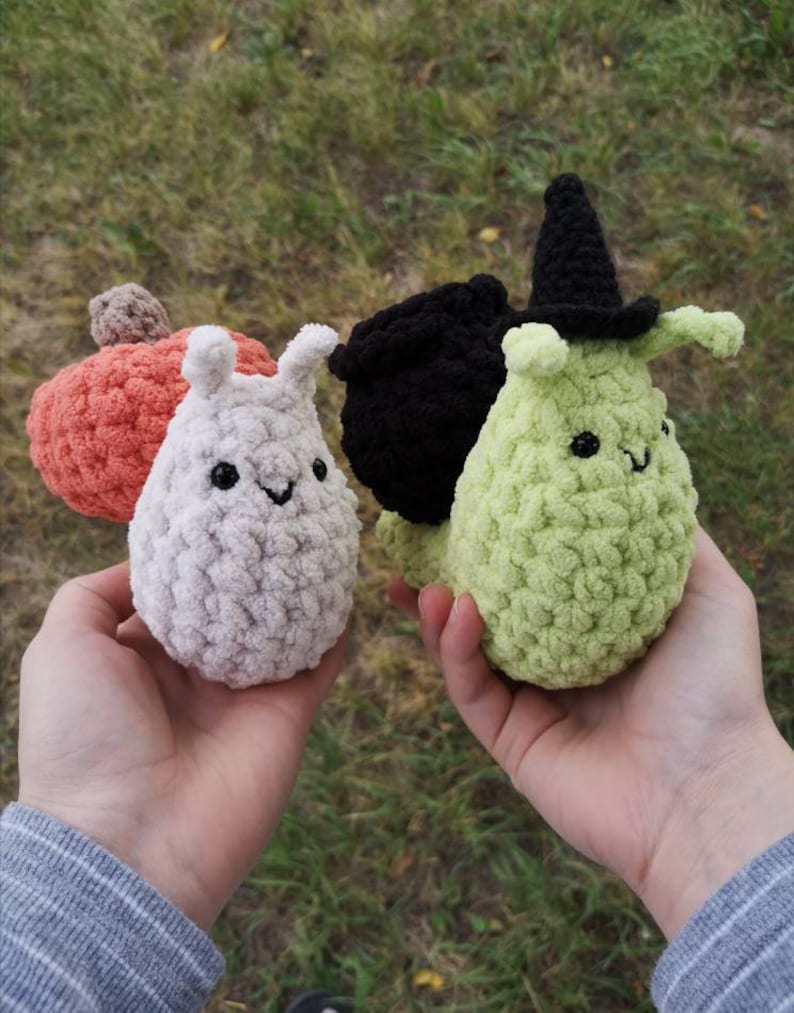 May include: Three crocheted plush toys in the shape of snails. The snails are white, green, and black. The green snail is wearing a black witch's hat.
