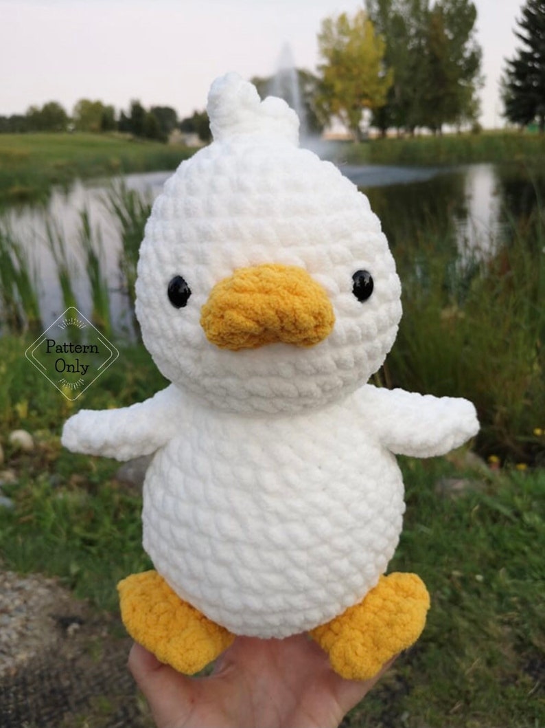 May include: White crocheted duck plush toy with yellow beak and feet. The duck is sitting on a hand.