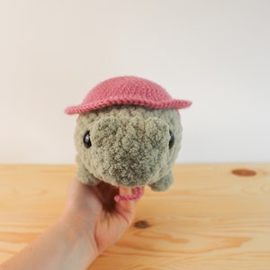 Op de afbeelding: Een gehaakt knuffeldier met een roze hoed. Het dier heeft een grijs lichaam en zwarte ogen. Het dier wordt vastgehouden door een hand.