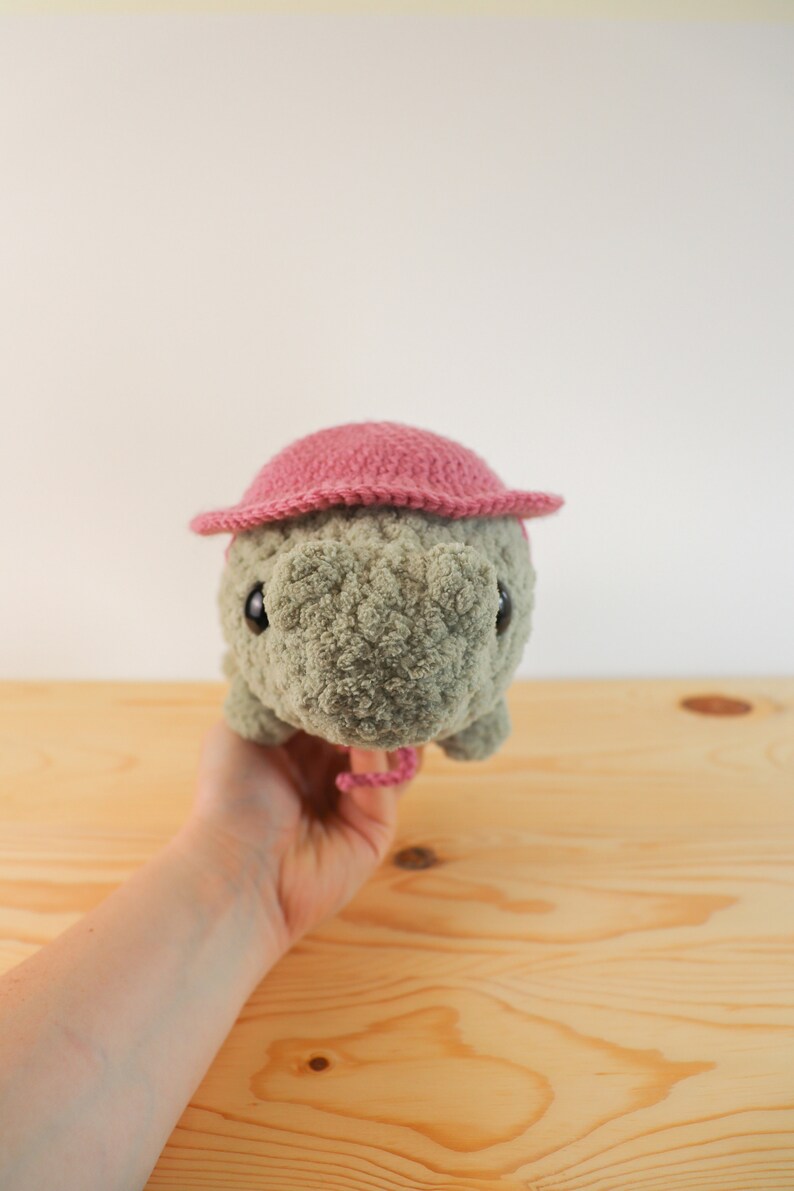 Op de afbeelding: Een gehaakt knuffeldier met een roze hoed. Het dier heeft een grijs lichaam en zwarte ogen. Het dier wordt vastgehouden door een hand.