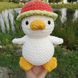 May include: White crocheted duck plush toy wearing a red and green crocheted watermelon hat. The duck has yellow feet and a yellow beak.