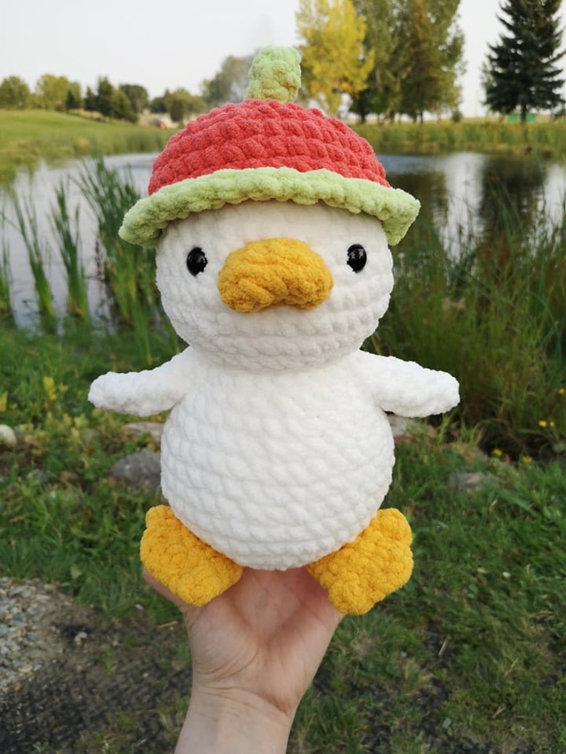May include: White crocheted duck plush toy wearing a red and green crocheted watermelon hat. The duck has yellow feet and a yellow beak.