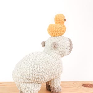 May include: A handmade, light gray crochet sheep with gray accents and a small, yellow crochet duck perched on its back. The sheep is standing on a wooden surface against a white background. The duck has a small red beak.
