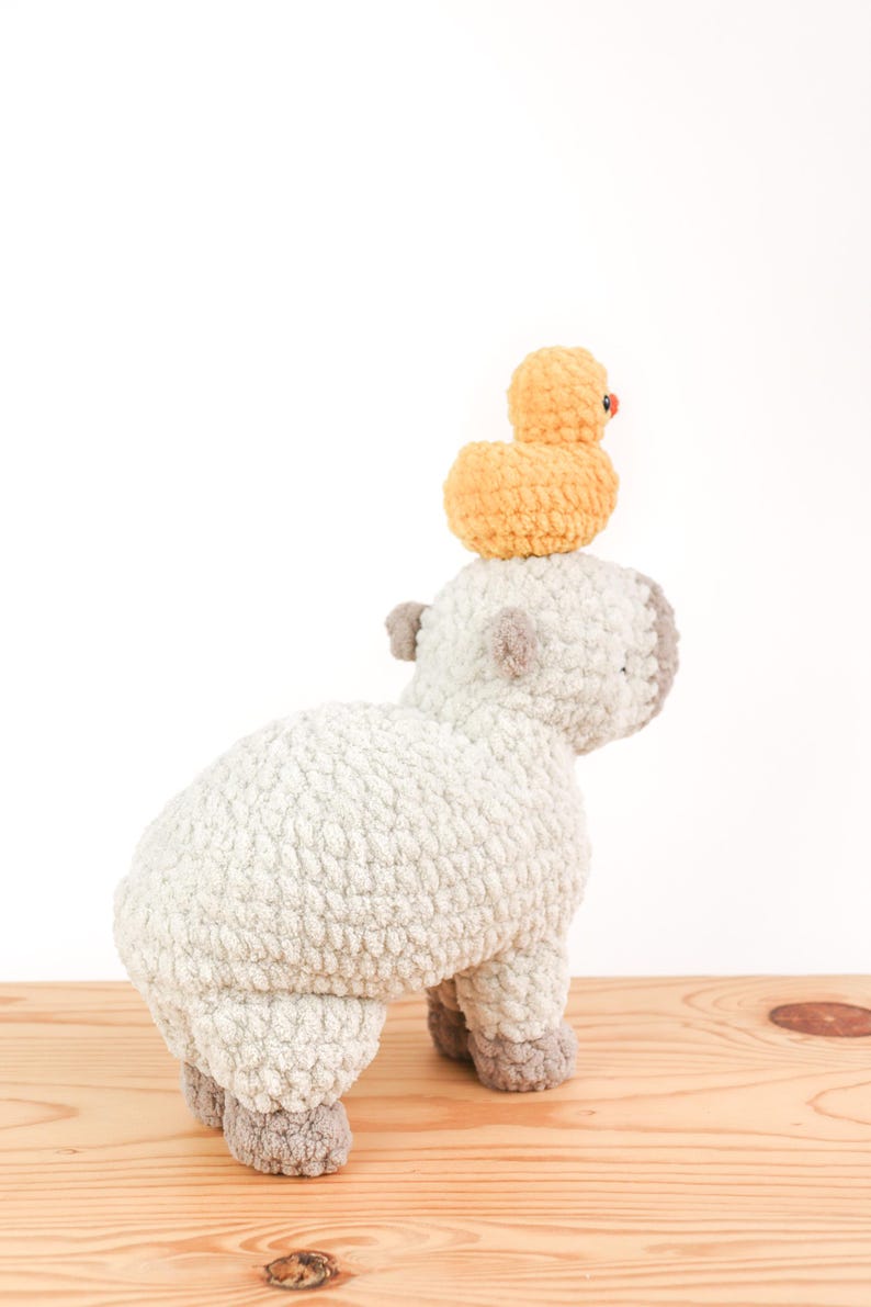 May include: A handmade, light gray crochet sheep with gray accents and a small, yellow crochet duck perched on its back. The sheep is standing on a wooden surface against a white background. The duck has a small red beak.
