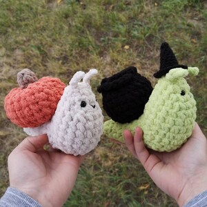 May include: Three crocheted snail plushies. The first snail is white with a pink pumpkin on its back. The second snail is green with a black witch's hat. The third snail is black with a black cauldron on its back.