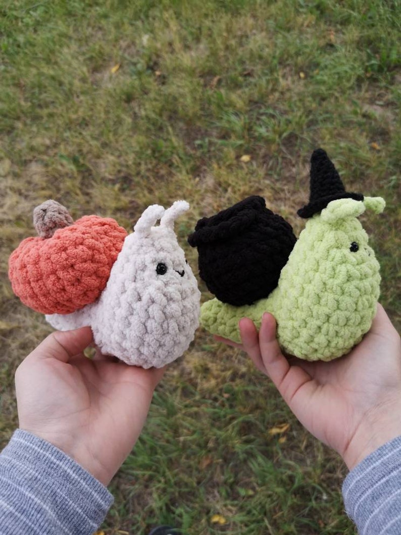 May include: Three crocheted snail plushies. The first snail is white with a pink pumpkin on its back. The second snail is green with a black witch's hat. The third snail is black with a black cauldron on its back.