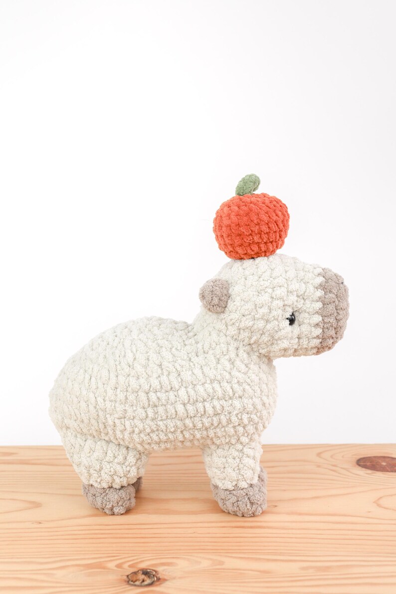 May include: A handmade, plush capybara with a small orange apple on its head. The capybara is light beige with gray accents on its face, ears, and feet. The apple has a green leaf. The toy is sitting on a wooden surface.
