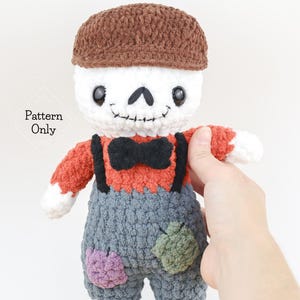 Może przedstawiać: Szydełkowa lalka szkieletora w brązowej czapce, pomarańczowej koszuli, szarych ogrodniczkach z łatkami i czarną muszką. Lalka ma białą głowę z czarnymi oczami i wyszywanym uśmiechem. Tekst "Pattern Only" znajduje się na zdjęciu.