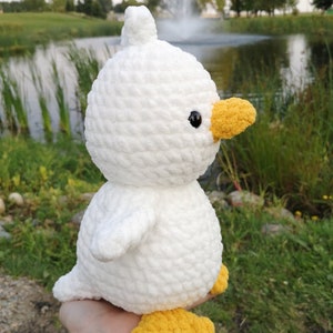May include: A white crocheted duck plush toy with yellow feet and beak. The duck is sitting on a hand.