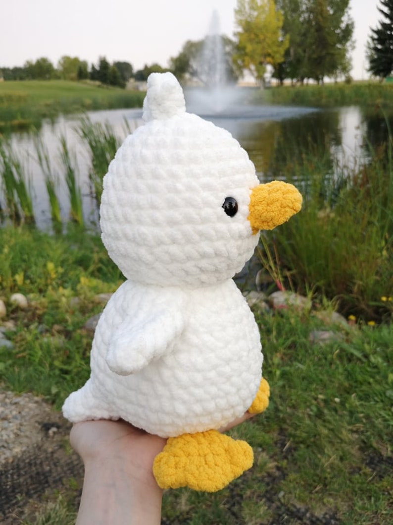 May include: A white crocheted duck plush toy with yellow feet and beak. The duck is sitting on a hand.
