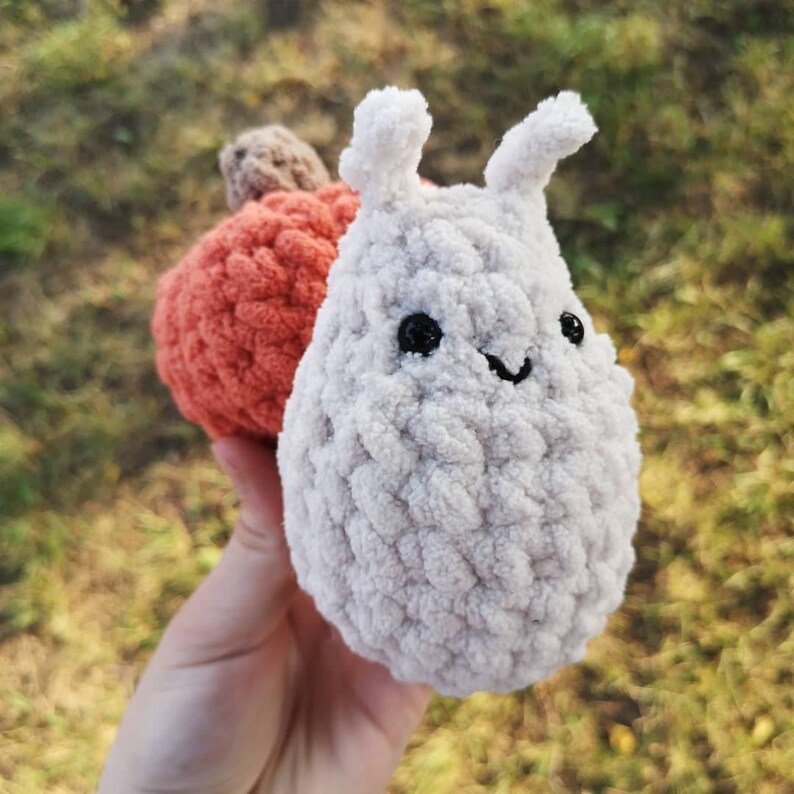 May include: A white crocheted plush toy shaped like a small animal with black eyes and a smiling mouth. The toy is sitting on top of a smaller orange crocheted pumpkin.