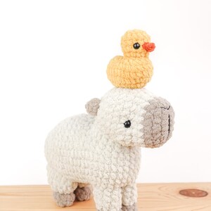 May include: A handmade crochet capybara toy in off-white with gray accents, topped with a small yellow duck with an orange beak. The plush toys are displayed on a light wooden surface against a white background.