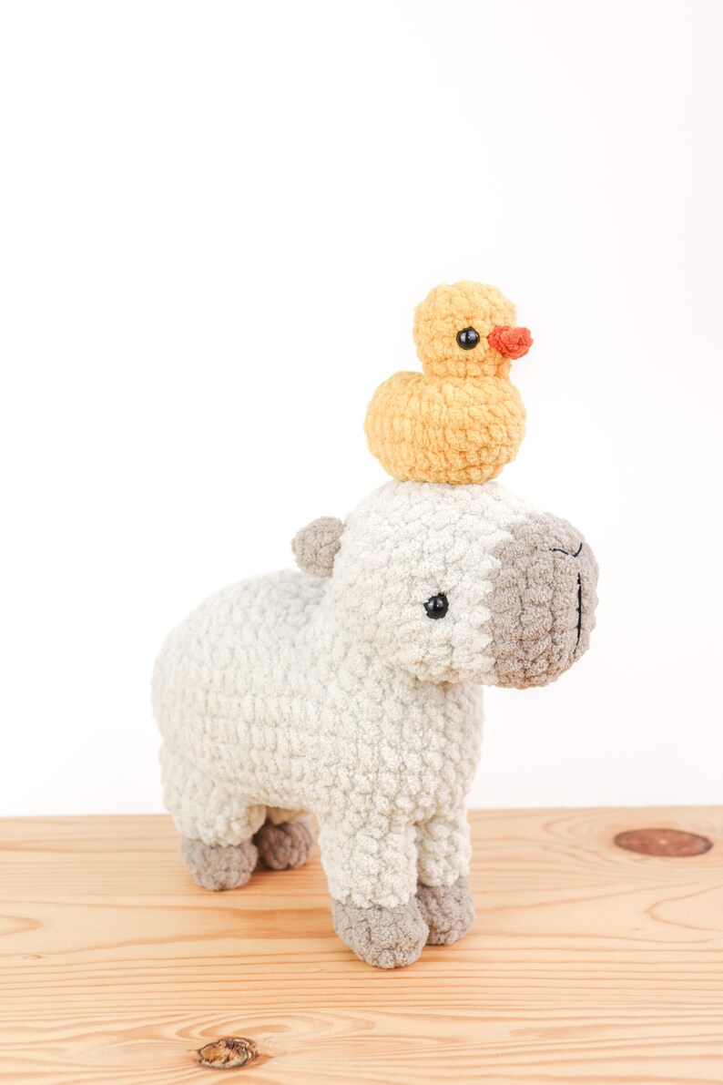 May include: A handmade crochet capybara toy in off-white with gray accents, topped with a small yellow duck with an orange beak. The plush toys are displayed on a light wooden surface against a white background.