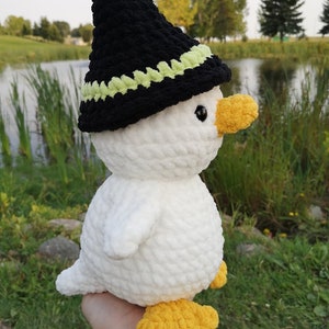 May include: A white crocheted duck wearing a black witch hat with a green trim. The duck has yellow feet and a yellow beak.
