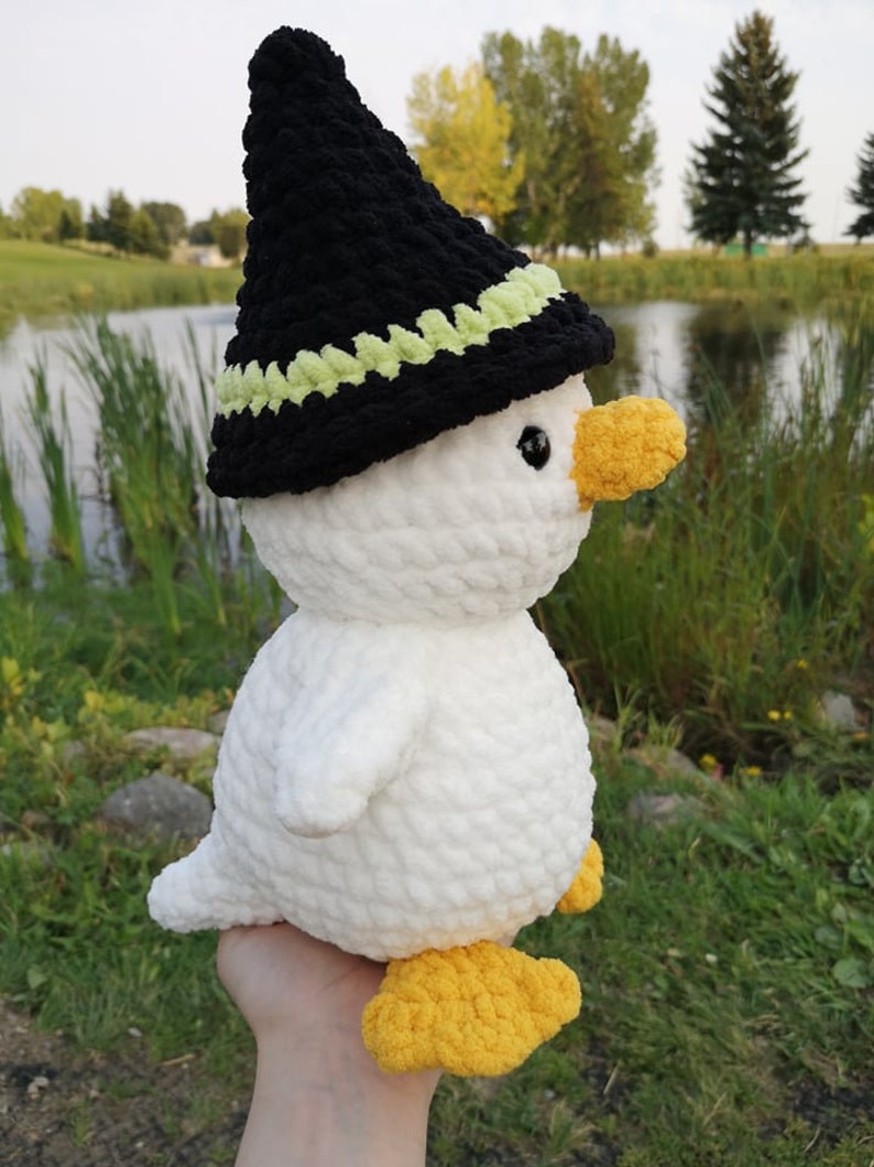 May include: A white crocheted duck wearing a black witch hat with a green trim. The duck has yellow feet and a yellow beak.