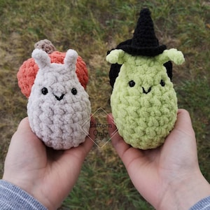 May include: Two crocheted plush toys, one is a white snail with an orange pumpkin shell and the other is a green witch snail with a black witch hat.  The toys are held in a person's hands.