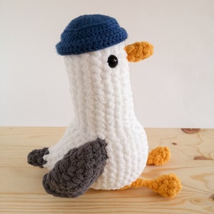 May include: A handmade white crochet seagull plush toy with a blue hat, gray wings, and orange beak and feet. The seagull is sitting on a wooden surface. The plush toy has black button eyes.