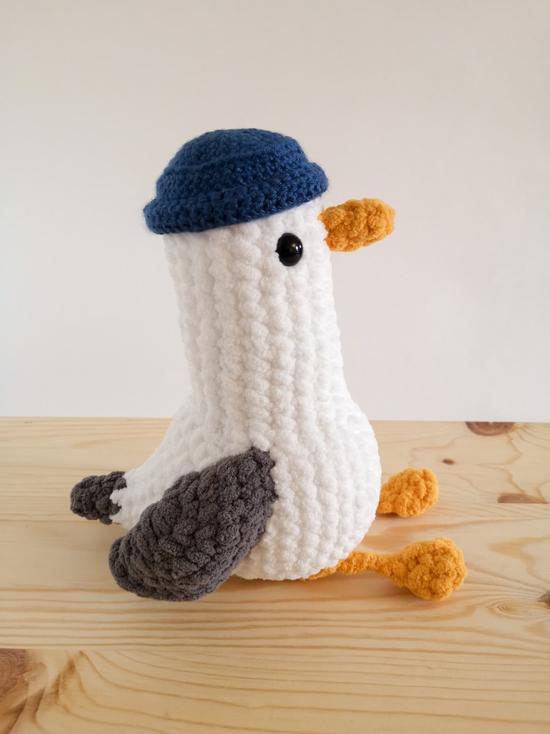 May include: A handmade white crochet seagull plush toy with a blue hat, gray wings, and orange beak and feet. The seagull is sitting on a wooden surface. The plush toy has black button eyes.